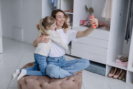 Young mother sits in a modern dressing room with a phone taking selfies with her twin daughtersの写真素材