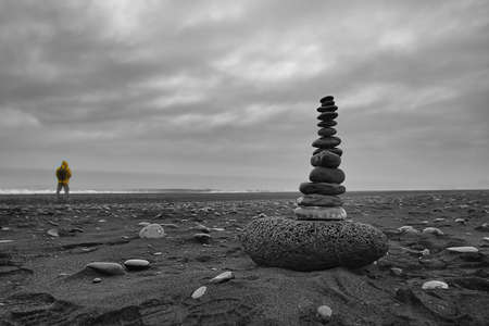 Small stone tower on a black beach in Iceland with a man in a yellow raincoat in the backgroundの写真素材