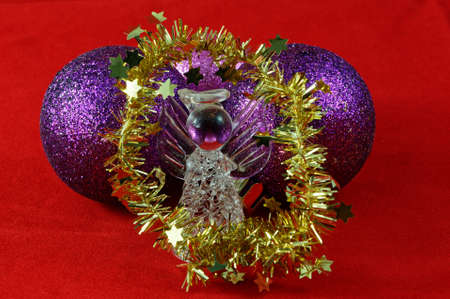 A glass Christmas angel is surrounded by gold tinsel and stands in front of purple baubles on a red backgroundの写真素材