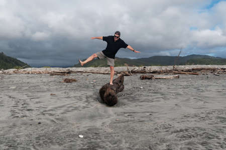 Testing his balance, an older man stands on one leg on a driftwood log on the beach, he's having funの写真素材