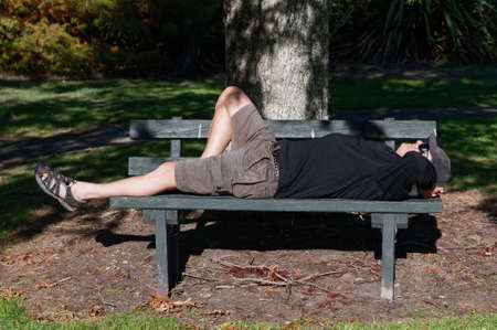 A man is lying on a park bench under the dappled light of a treeの写真素材
