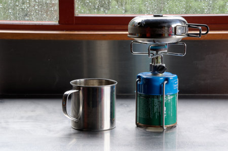 A small stainless steel kettle with folding handles is heating on top of the portable gas burner, the stainless steel mug is waiting near byの写真素材