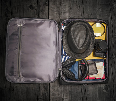 Open traveler's bag with clothing, accessories, money and passport on wooden floor background....