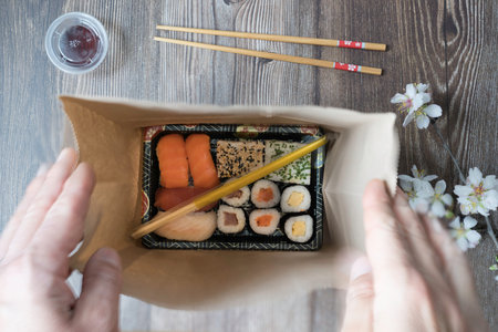 Top view of a take-away box with sushi rolls and chopsticks in a brown recyclable paper bag. Food...