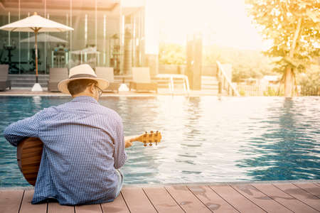 Romantic young man sitting on the pool at sunset with playing the guitarの写真素材