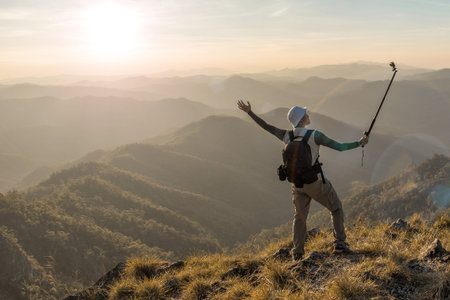 Photographer with action camera in hand on top of mountain. Hiker climbed on peak of rock above foggy valley.の写真素材