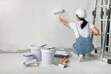 girl paints a white wall with a roller. Repair of the interior. Young female decorator painting a wall in the empty room, concept builder or painter in helmet with paint roller over the empty room.の写真素材