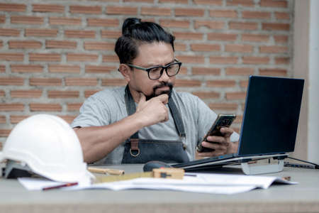 Male architect or engineer working at home drawing the construction project, An architect working on a laptop using a mobile phone on the working desk. Business and Technology concept.の写真素材