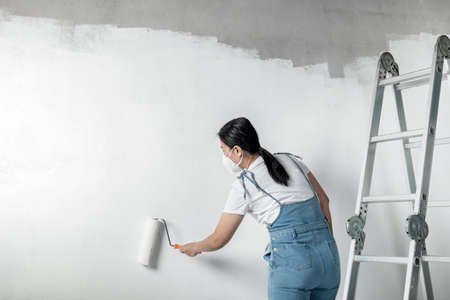 girl paints a white wall with a roller. Repair of the interior. Young female decorator painting a wall in the empty room, concept builder or painter in helmet with paint roller over the empty room.の写真素材