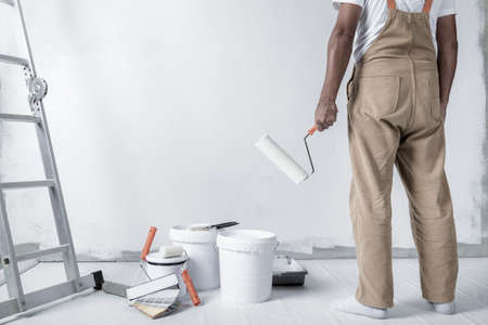 a man paints a white wall with a roller. Repair of the interior. Young male decorator painting a wall in the empty room, concept builder or painter in helmet with paint roller over the empty room.の写真素材