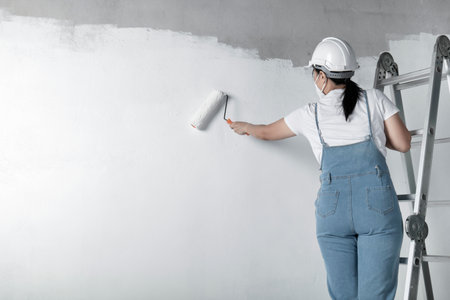 girl paints a white wall with a roller. Repair of the interior. Young female decorator painting a wall in the empty room, concept builder or painter in helmet with paint roller over the empty room.の写真素材