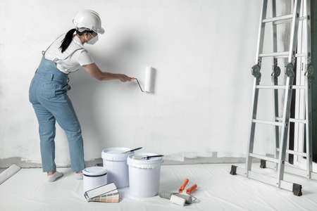girl paints a white wall with a roller. Repair of the interior. Young female decorator painting a wall in the empty room, concept builder or painter in helmet with paint roller over the empty room.の写真素材