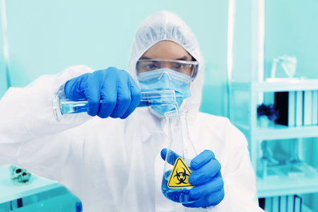 Male Scientist in PPE Suit working in Lab while Pouring Solution from Cylinder to Erlenmeyer Flask with BioHazard Sign. Blue Tone. SARS-CoV-2 , Covid-19 THEME.の写真素材