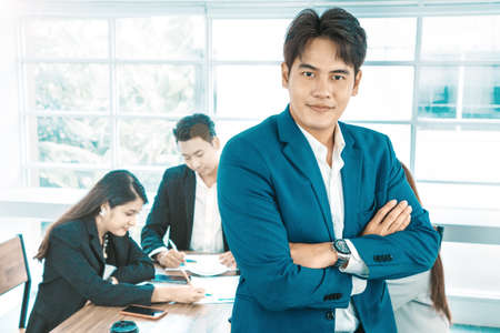 Portrait of a handsome CEO smiling, Portrait of a positive looking young business professional standing with his arms crossed with coworkers talking in the background.の写真素材