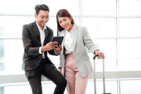 Business couple standing together with baggage and checking flight time on tablet near the window at the departure area at the airportの写真素材