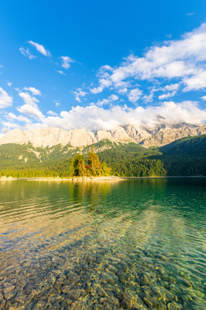 Eibsee Lake, Hill Slopes with Forest, Island and Zugspitze Mountain on Sunny Evening. Ripple on...