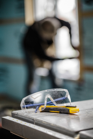 Protective Glasses and Utility Knife on a Table Saw with a worker in backgroundの写真素材
