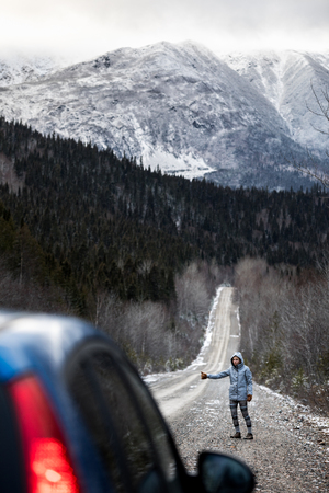Lonely Young Woman Hitchhiking on a Winter Road with Beautiful Snowy Mountains in Backgroundの写真素材