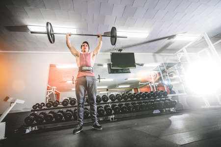 From below view of sportive man in gym lifting heavy barbell above headï¿½doing military press.の写真素材