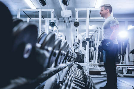 Side view of bodybuilder lifting barbell near rows of metal dumbbell in gym.の写真素材