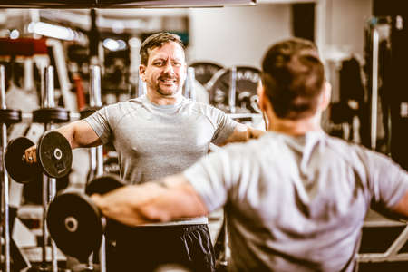 Back view of brutal sportsman raising dumbbells laterally training in front of mirror in gym.の写真素材