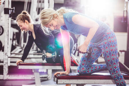 Side view of smiling women doing dumbbell row on benches in modern gym.の写真素材