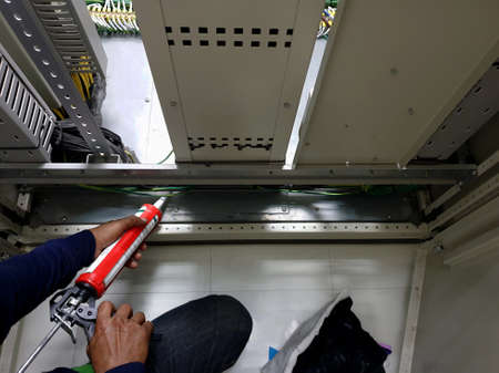 A worker uses silicone adhesive with a glue gun to seal the floor of the electrical control cubicle.の写真素材