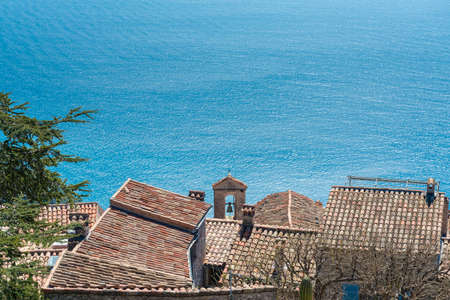 View down to the azure blue Mediterranean sea over the terracotta rooftops of Eze Village on a sunny dayの写真素材