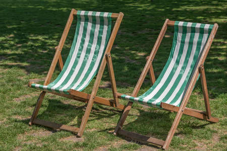 A pair of empty traditional green striped wooden deckchairs in a park in the summer sunの写真素材