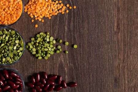 three types of legumes, beautifully laid out in cups and on a wooden background - red beans, green peas and orange lentils. Top view. Vegetarian food. Unprepared.の写真素材