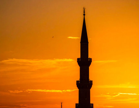 Dramatic mosque minaret stands silhouetted against a vibrant orange sunset sky evoking a sense of peace and spirituality Ideal for religious or travel themes.の素材