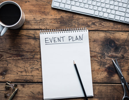 An event plan notebook lies open with a pencil, coffee, keyboard, and scissors on wood.の素材