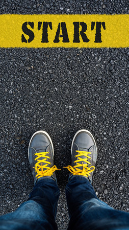 Feet wearing gray sneakers with yellow laces stand at a yellow start line on asphalt.の素材