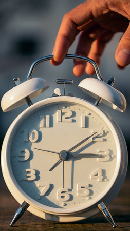 A hand reaches to turn off a white analog alarm clock with two metal bells on top.の素材
