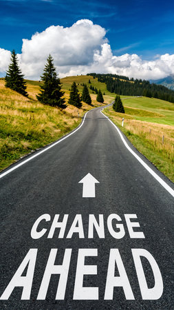 A winding asphalt road with "Change Ahead" painted on it leads through green rolling hills under a blue sky.の素材