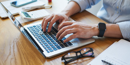 A person's hands typing on a laptop with a calculator, glasses, and phone on a wooden desk.の素材