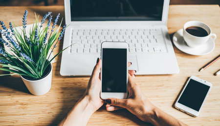 Hands holding a smartphone with a laptop, coffee and plant on a wooden desk. Clear details and vibrant colors...の素材