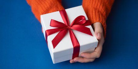 Female hands holding white gift box with red ribbon on blue background.の素材