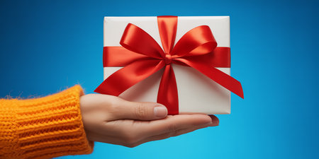 Female hand holding a white gift box with red ribbon on blue backgroundの素材