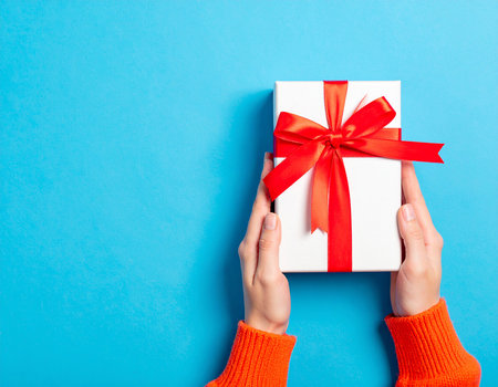 Female hands holding white gift box with red ribbon on blue background.の素材