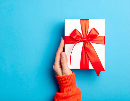 Female hand holding a white gift box with red ribbon on blue backgroundの素材