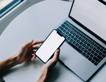 Mockup image of woman's hands holding smart phone with blank white screen and laptop on white tableの素材
