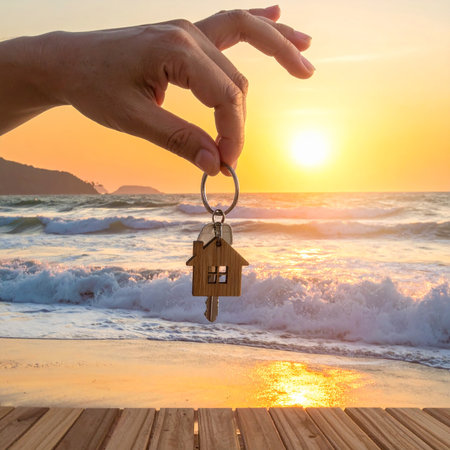 Hand holding key with house shaped keychain over blurred sea and sunset backgroundの素材