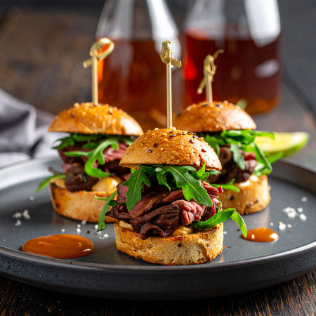 Three gourmet beef sliders with arugula and sauce are presented on a dark plate. Clear details and vibrant co...の素材