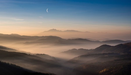 Misty mountains at sunrise with a crescent moon in a clear blue sky. Clear details and vibrant colors enhance...の素材