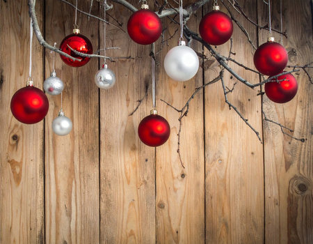 Red and silver Christmas ornaments hang from bare branches against a rustic wooden wall.の素材