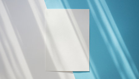 A blank white paper sheet rests on a split blue and white background with dappled light shadows.の素材