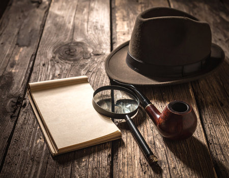 Detective's fedora hat, pipe, magnifying glass, and notebook on a rustic wooden surface.の素材
