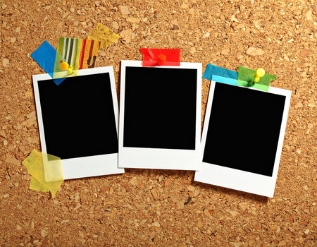 Three blank instant photo frames attached to a corkboard with colorful tapes and pins.の素材
