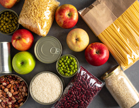 Overhead view of canned goods, pasta, rice, beans, and fresh apples arranged on a gray surface.の素材